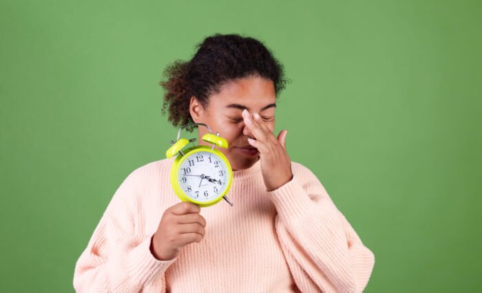 Beautiful african american woman on green background with alarm clock sleepy tired exhausted yawning