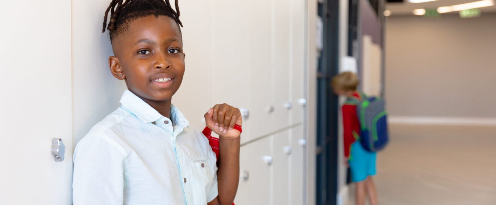 Portrait of smiling african american elementary schoolboy standing by locker in school. unaltered, childhood, education and back to school concept.